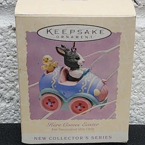 May include: Keepsake ornament box featuring a bunny and chick in a blue car with red wheels. The box has the text "Here Comes Easter" and "New Collector's Series".