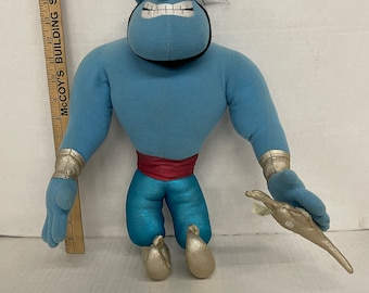 May include: Blue plush Genie from Disney's Aladdin, with a mischievous expression, wearing a red sash and gold pants. He is holding a gold staff in his right hand.