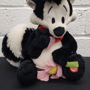 May include: A plush skunk toy with black and white fur, a red bow tie, and a pink flower. The toy has the text "PRESS HERE" on its foot. The skunk is sitting on a black surface.