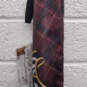 May include: A closed, maroon and black plaid umbrella with a black handle and strap. The umbrella has a tag that reads "Harry Potter" and features a crest. The umbrella is against a white brick wall.