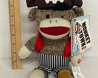 May include: A brown and white sock monkey wearing a brown pirate hat with a white skull and crossbones design. The monkey is wearing a red scarf and black and white striped pants. The monkey is sitting on a white surface. The tag on the monkey reads "Monkey Town USA Try Me!"