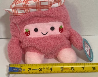 May include: A pink plush toy shaped like a rectangle with a smiling face, two small strawberries, and a plaid hat. The toy has small arms and legs. The toy measures approximately 4 inches in length.
