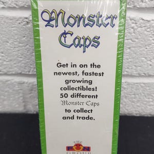 May include: A sealed box of "Monster Caps" collectibles. The box features the title in a stylized font with green and yellow accents. The text promotes the collection of 50 different Monster Caps for trading.
