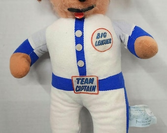 May include: A plush toy dog wearing a blue baseball cap, white and blue baseball uniform, and blue shoes. The uniform has the text "Big Leaguer" and "Team Captain" on it.
