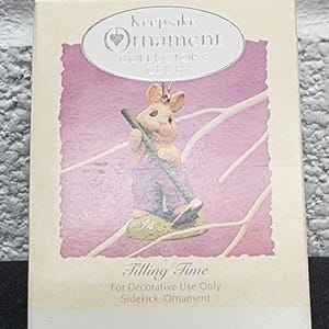 May include: A decorative ornament box featuring a bunny figurine holding a green pole. The box has the text "Keepsake Ornament Collector's Club" and "Tilling Time For Decorative Use Only Sidekick Ornament."