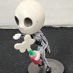 May include: A black and white striped figure of Jack Skellington from the Nightmare Before Christmas movie holding a red and green bottle with a white butterfly on top. The figure is standing on a black base with the word "Bottle" written on it.