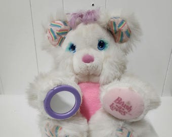 May include: White plush teddy bear with rainbow details on its ears and paws. The bear has blue eyes, a pink nose, and a pink heart on its chest. The bear is holding a purple mirror in its left paw. The bear's right paw has the text "Brush 'n' Love" printed on it.
