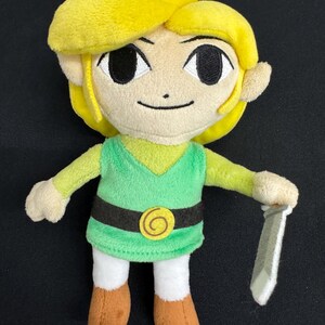 May include: A plush toy of a character with yellow hair, green tunic, and brown boots, holding a silver sword. The character has a smiling face with large eyes and is wearing a black belt with a gold buckle. The toy is approximately 8 inches tall.
