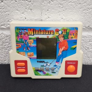 May include: Electronic miniature golf game by Tiger. The handheld game features a screen, direction and swing/hit buttons, and illustrations of golfers. The game is white with orange and red accents.