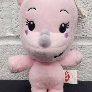 May include: A pink plush rhino toy with a gray horn, black eyes, and eyelashes. The toy has a tag that reads "Lulu". The rhino is standing on a black surface.