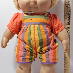 May include: A vintage doll with blonde yarn hair and blue eyes, wearing an orange short-sleeved shirt and striped overalls in shades of orange, yellow, green, and purple. The doll is approximately 12 inches tall.
