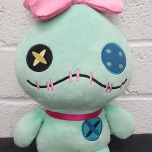 K&ouml;nnte beinhalten: Eine mintgr&uuml;ne Pl&uuml;schpuppe mit einer gro&szlig;en rosa Satinschleife oben und einer gelben Haartolle. Die Puppe hat ein schwarzes Auge mit einem gelben Kreuz, ein blaues Knopfauge, einen gestickten Mund, ein rosa Band als Halsband und einen blauen Knopf.