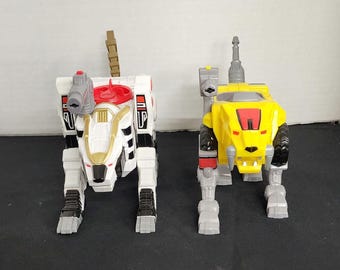 May include: Two toy robot lions, one white with red and gold accents and one yellow with silver accents. Both have a similar design with a head, body, and legs.