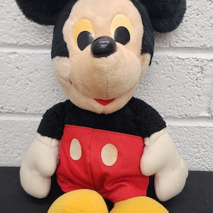 May include: A plush Mickey Mouse toy with black ears, a black body, white gloves, red shorts, and yellow shoes. The toy has a tan face with black eyes and a red smile. A classic Disney character.