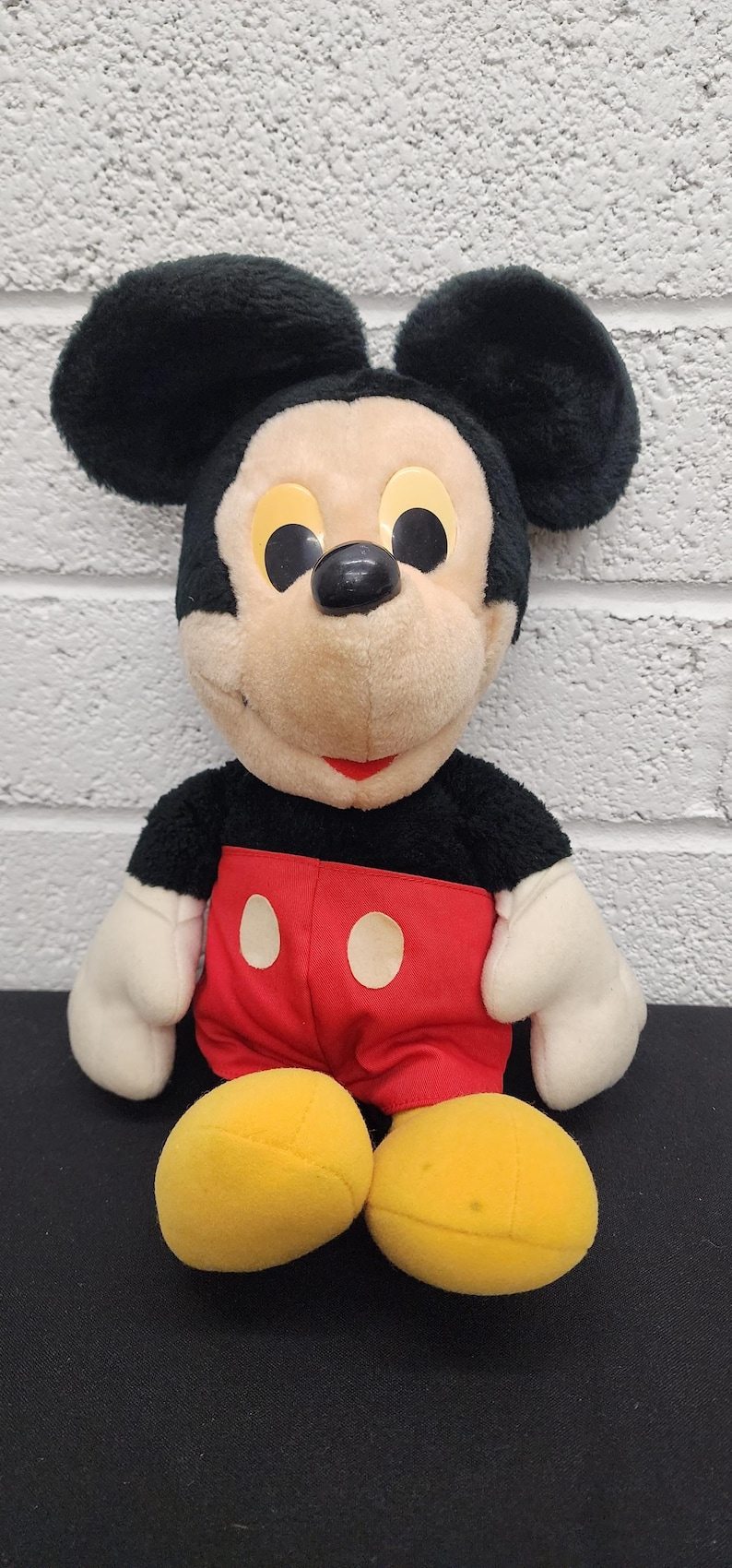 May include: A plush Mickey Mouse toy with black ears, a black body, white gloves, red shorts, and yellow shoes. The toy has a tan face with black eyes and a red smile. A classic Disney character.