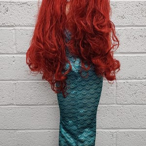 May include: A mermaid costume featuring a teal scaled bodice and a flowing, light blue tail. The costume includes a long, vibrant red wig. The outfit is designed to resemble a mermaid.