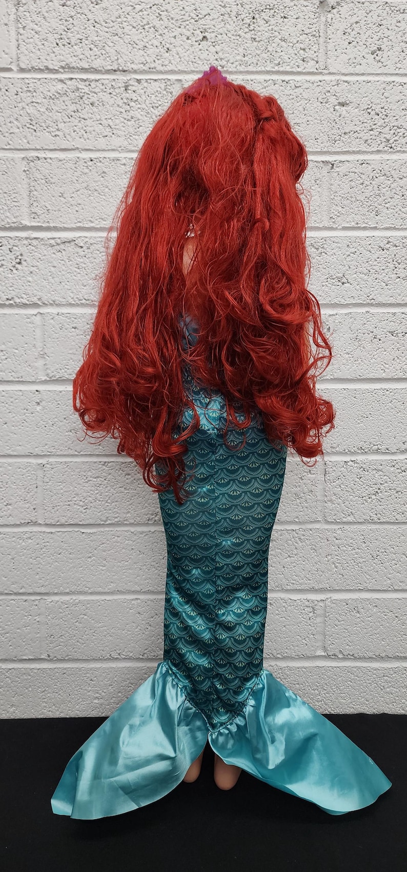 May include: A mermaid costume featuring a teal scaled bodice and a flowing, light blue tail. The costume includes a long, vibrant red wig. The outfit is designed to resemble a mermaid.