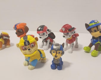 May include: A collection of Paw Patrol character figurines. The figures are of various colors, including yellow, blue, red, and brown. Each figure is wearing a uniform and hat. A wooden ruler is in the background.