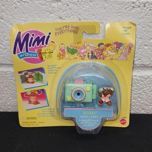 May include: A vintage Mimi the Good toy set featuring a mint green camera and a small bear figure named Kayo. The packaging is yellow with illustrations and text that reads "Kayo and his camera".