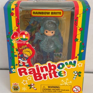 May include: A small plastic doll of Rainbow Brite in a blue winter outfit with a white fur trim. The doll is packaged in a yellow box with a red, orange, yellow, green, blue, indigo, and violet striped border. The box has a picture of Rainbow Brite on the front and the words "Rainbow Brite" and "40 Years of" in large letters. The box also has a warning label that says "CHOKING HAZARD - Small parts. Not for children under 3 years."