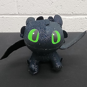 May include: A plush toy of a black dragon with large green eyes and small black wings. The dragon has a textured, sparkly appearance and is sitting on a black surface.