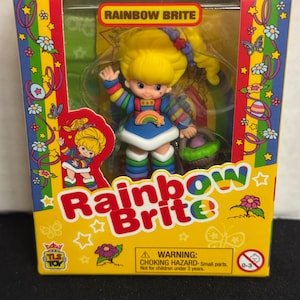 May include: A Rainbow Brite doll in a box. The doll has yellow hair, a blue dress, and rainbow accents. The box is yellow, red, and green with the words "Rainbow Brite" and a warning about small parts.