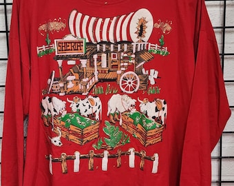 May include: Red long-sleeve shirt with a Western-themed graphic. The graphic includes a sheriff's office, cows, and a stagecoach. The shirt has a crew neck and is decorated with small studs around the collar.