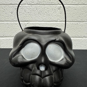 May include: Black plastic skull-shaped Halloween candy bucket with a black metal handle. The skull has white eye sockets and a white nose. The bucket is designed to resemble a human skull.