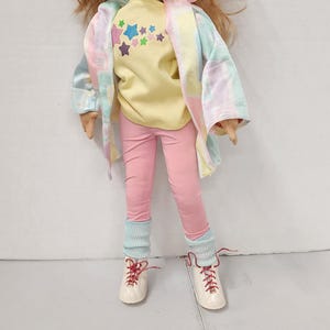 May include: A doll with long brown hair wearing a yellow shirt with a rainbow star design, a pink tie-dye jacket, pink pants, and white shoes with red laces.