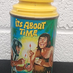 May include: Vintage metal lunchbox with a yellow lid and base. The lunchbox features a cartoon illustration of two cavemen holding matches, with the text "Its About Time" in yellow letters.