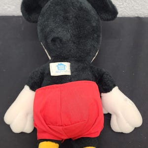 May include: A plush Mickey Mouse doll, viewed from the back, featuring a black body, ears, and head. It has white gloves, red shorts, and yellow shoes. A tag is visible on the back. The doll is on a black surface.