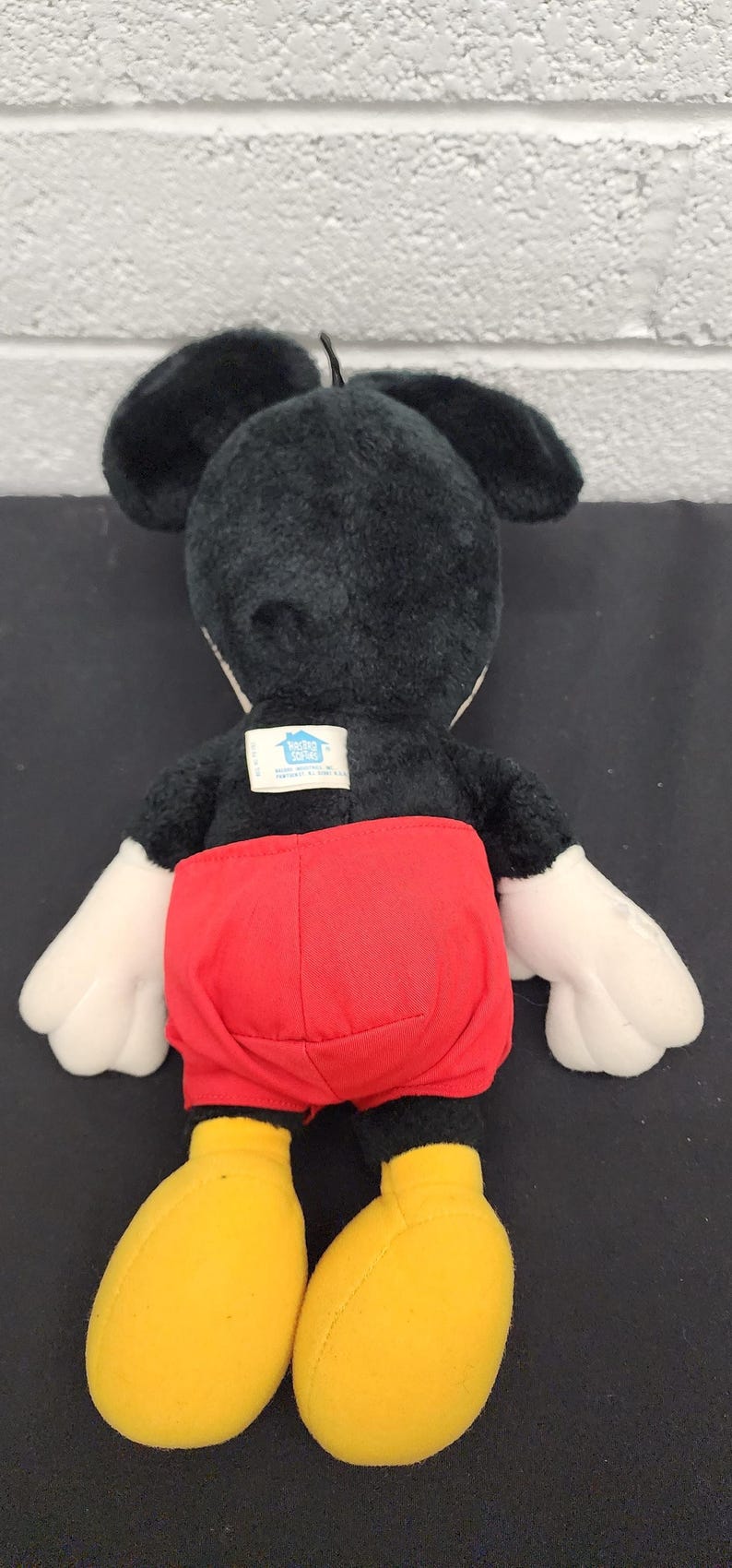 May include: A plush Mickey Mouse doll, viewed from the back, featuring a black body, ears, and head. It has white gloves, red shorts, and yellow shoes. A tag is visible on the back. The doll is on a black surface.