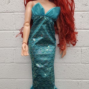 May include: A large doll of a mermaid with long red hair and blue eyes. She wears a teal top and a mermaid tail with a scale pattern. The doll is approximately 30 inches tall.