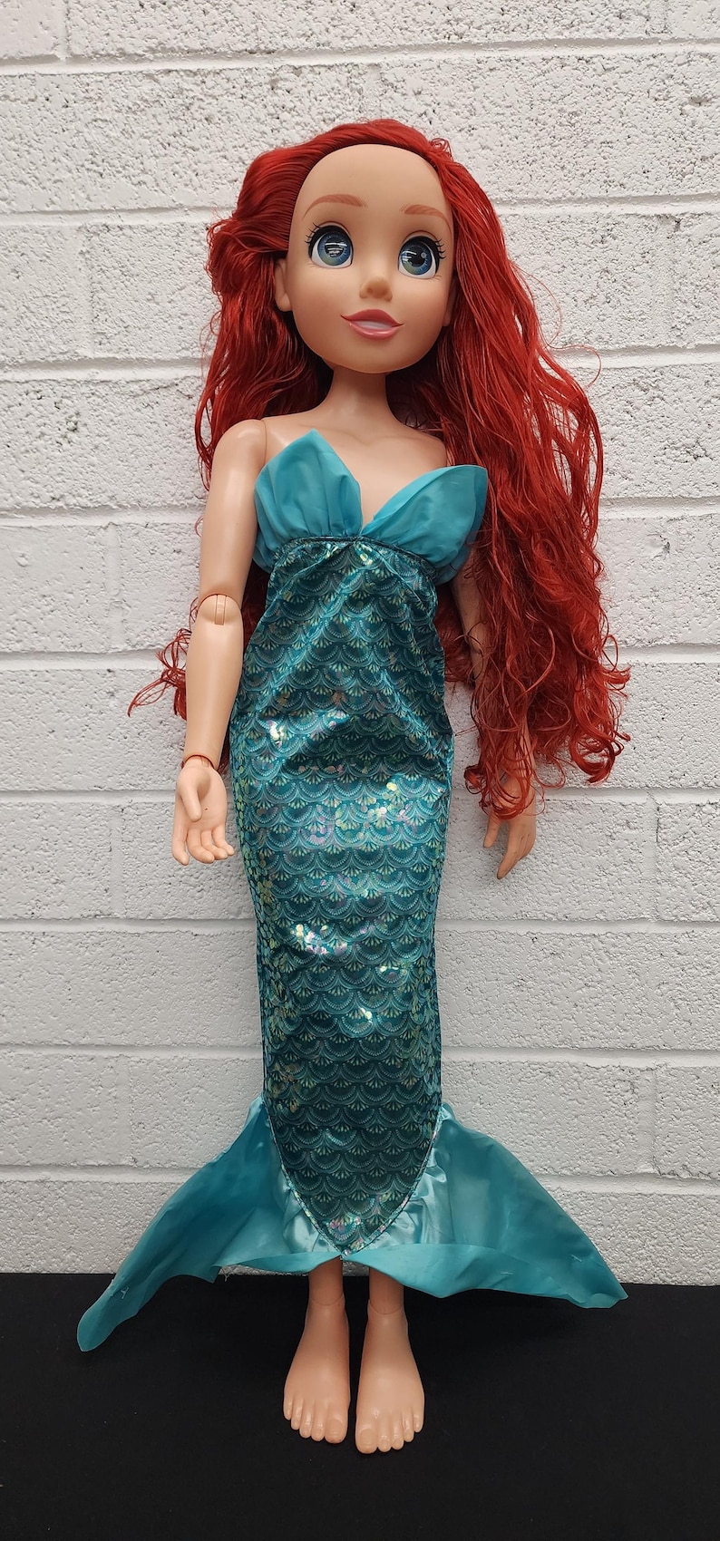 May include: A large doll of a mermaid with long red hair and blue eyes. She wears a teal top and a mermaid tail with a scale pattern. The doll is approximately 30 inches tall.