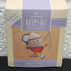 May include: Hallmark Keepsake Ornament box featuring an egg character playing tennis. The box is white and purple with the text "Collector's Series" and "Eggs in Sports". The ornament is for decorative use only.