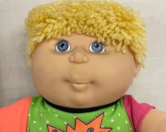 May include: A Cabbage Patch Kid doll with blonde, curly hair and blue eyes. The doll wears a green shirt with white polka dots and the word "SWAK" in an orange burst design. The sleeves are pink and orange.