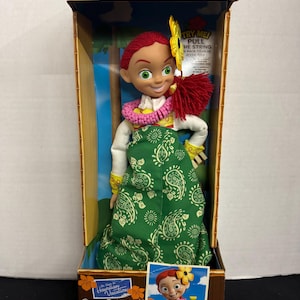 May include: A Disney Store Special Edition Toy Story Hawaiian Talking Jessie doll in a box. Jessie has red hair, a flower, and a lei. She wears a green paisley dress and a white shirt. The box says "Try Me! Pull the string back to hear Jessie say..."