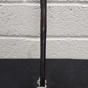 May include: A dark brown walking stick with a stylized animal head handle. The handle is dark and the base is a detailed silver face. The stick is standing on a black surface against a white brick wall.