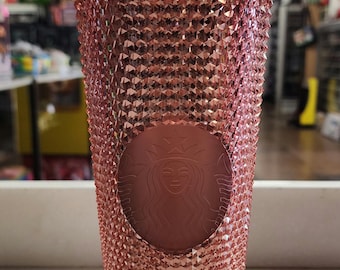 May include: A pink reusable Starbucks cup with a diamond-like texture and a clear plastic straw. The cup has the Starbucks logo embossed on the front.