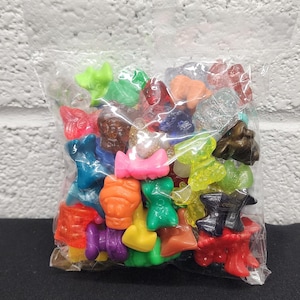 May include: A clear plastic bag filled with a collection of small, colorful figurines. The figurines are various shapes and colors, including red, blue, green, yellow, and orange. The background is a white brick wall.
