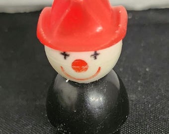 May include: A red and white plastic toy figurine of a clown wearing a red firefighter hat. The clown has a black smile and black eyes.