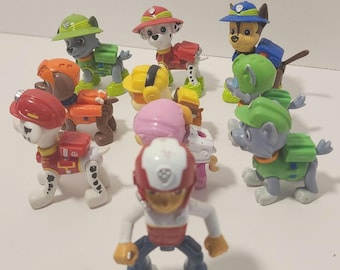 May include: A collection of colorful Paw Patrol character figurines. The figures are in various poses and outfits, including helmets and vests. The characters are primarily dogs, with different colored uniforms and accessories.