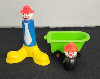 May include: Two plastic clown figurines with red hats and white faces. One clown is standing on yellow shoes and has a blue body with a white tie. The other clown is in a green wagon with black wheels.