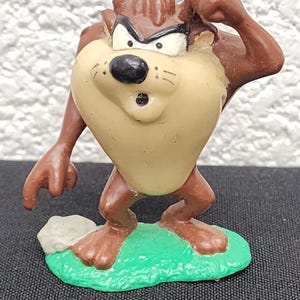 May include: A Taz figurine, a character from Looney Tunes, stands on a green base. The figure is brown and tan with a stern expression. The figurine is approximately 3 inches tall.