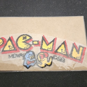 May include: A Pac-Man patch on a cardboard backing. The patch features the Pac-Man logo in yellow and red, along with images of Pac-Man and ghosts. The word "MIDWAY" is also visible.