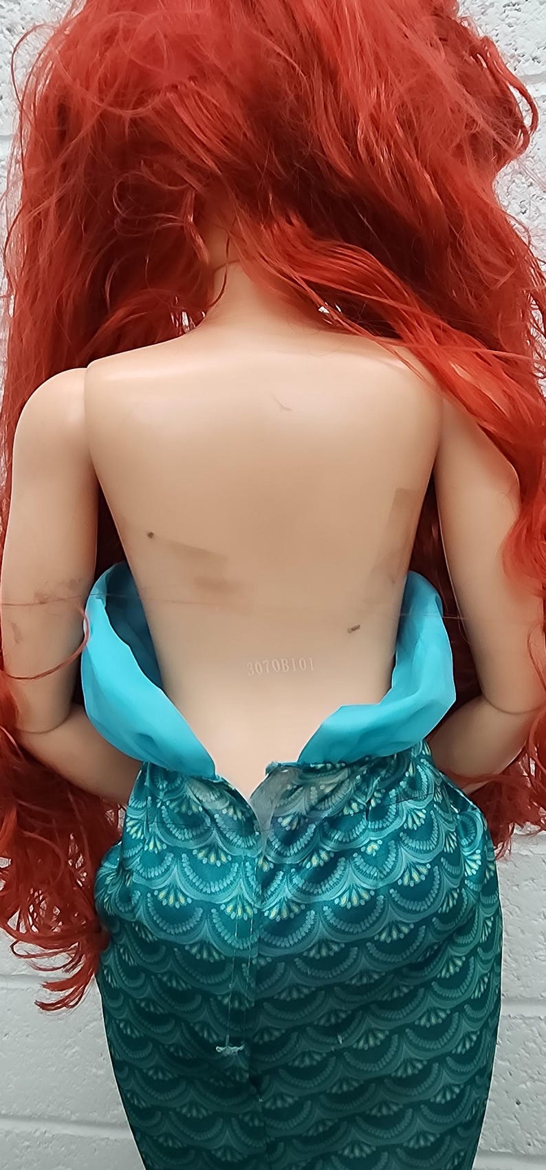 May include: A doll with long, vibrant red hair and a teal mermaid tail. The doll's torso is visible, with a teal top and a patterned skirt. The doll's back is visible, with the number "30708101" printed on it.