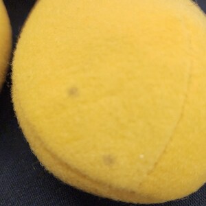 May include: Close-up of a yellow plush toy's foot, with a black and red head visible in the background. The foot has a few small brown spots. The toy is on a dark blue fabric surface.