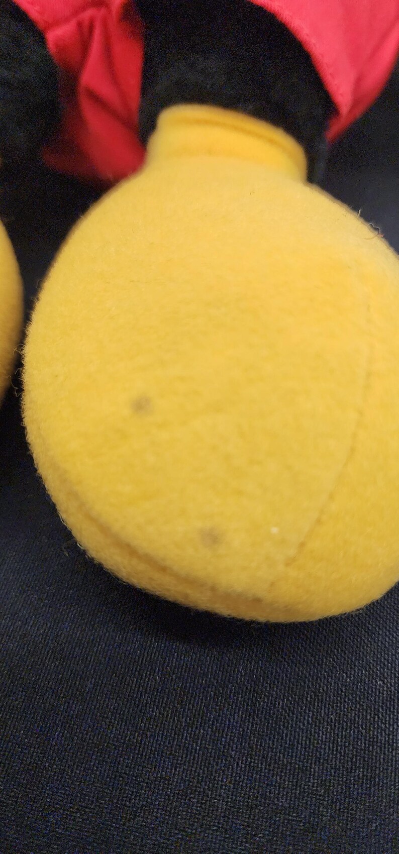 May include: Close-up of a yellow plush toy's foot, with a black and red head visible in the background. The foot has a few small brown spots. The toy is on a dark blue fabric surface.