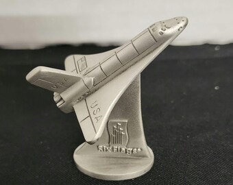 May include: A silver-colored pewter figurine of a space shuttle with the words "USA" and "Six Flags" on the base.
