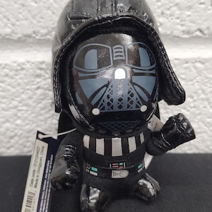 May include: A plush Darth Vader figure with a black helmet and suit. The figure has a detailed face and chest plate design. The Star Wars logo is visible on a tag.